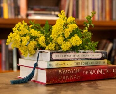 A stack of three books with a bookmark in the top book. The pile has a bunch of yellow flowers on top.